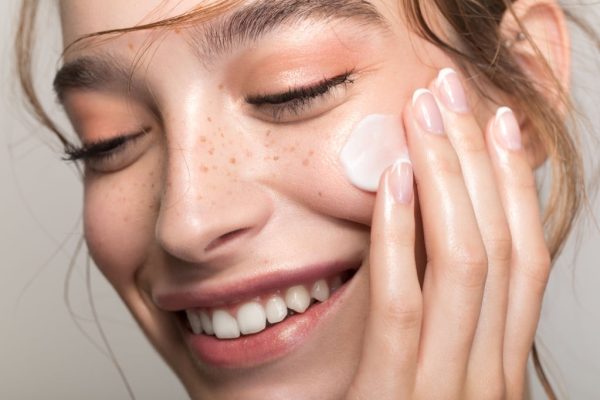 Closeup studio shot of a beautiful young woman with freckles skin, applying moisturiser to her face. Posing against a grey background